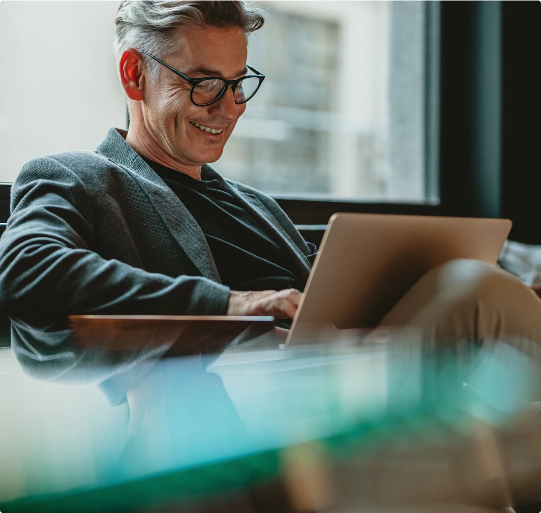 Photo of a man typing on a laptop and smiling while sitting on a couch.